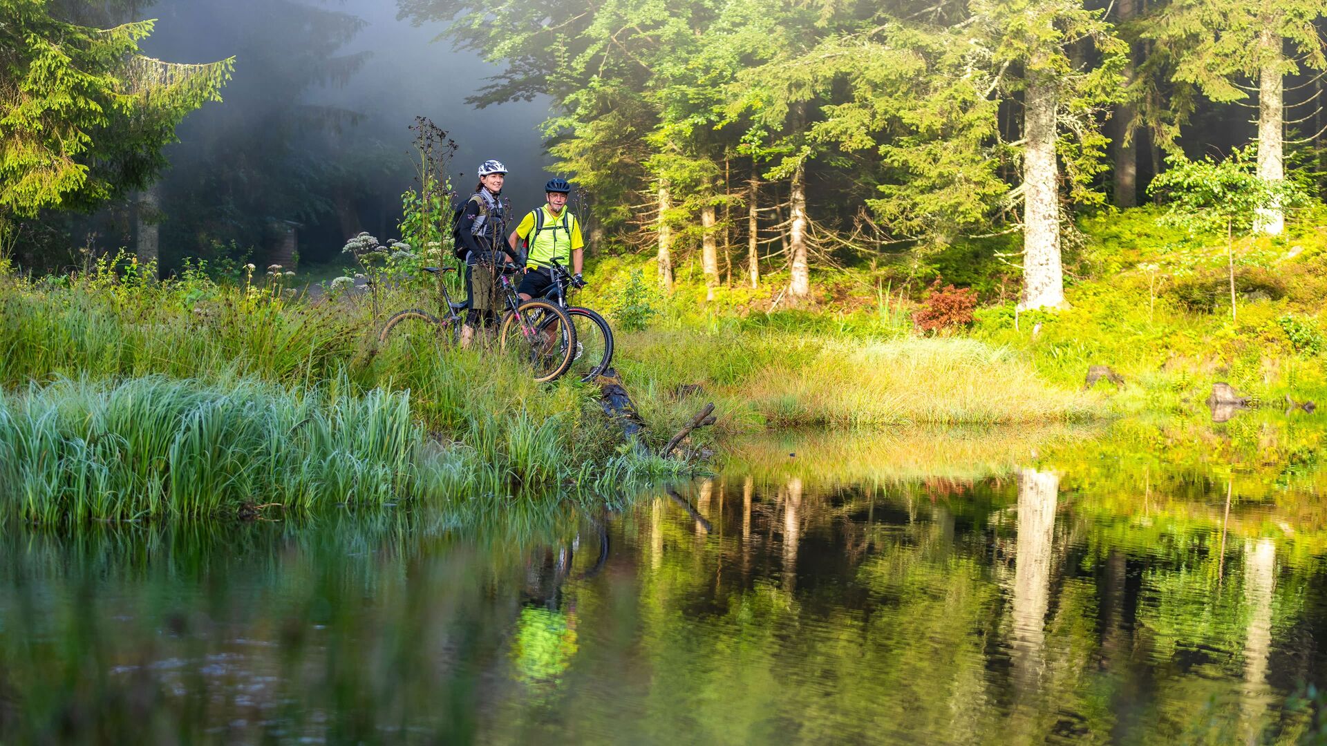 Zwei Radfahrer stehen mit Mountainbikes an einem idyllischen Waldsee in der Natur rund um den Gasthof Kammbräu